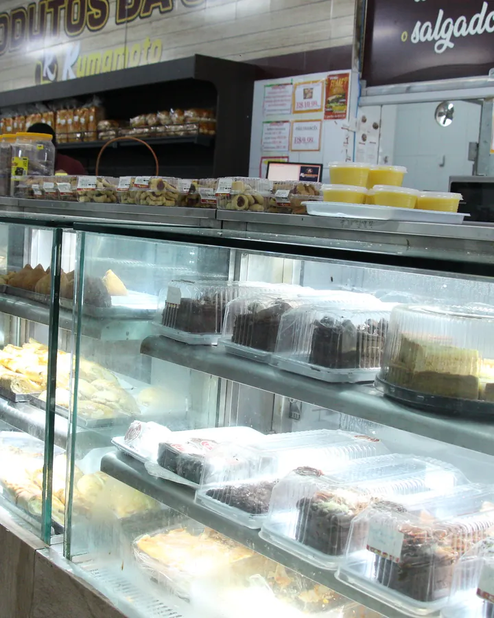 Balcão da confeitaria do Kumamoto com bolos, doces e salgados sob a placa Produtos da Casa