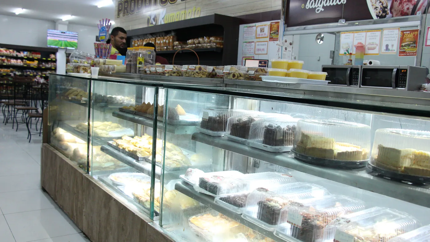 Balcão de vidro da confeitaria do Kumamoto, com bolos e tortas em exposição e a placa Produtos da Casa ao fundo